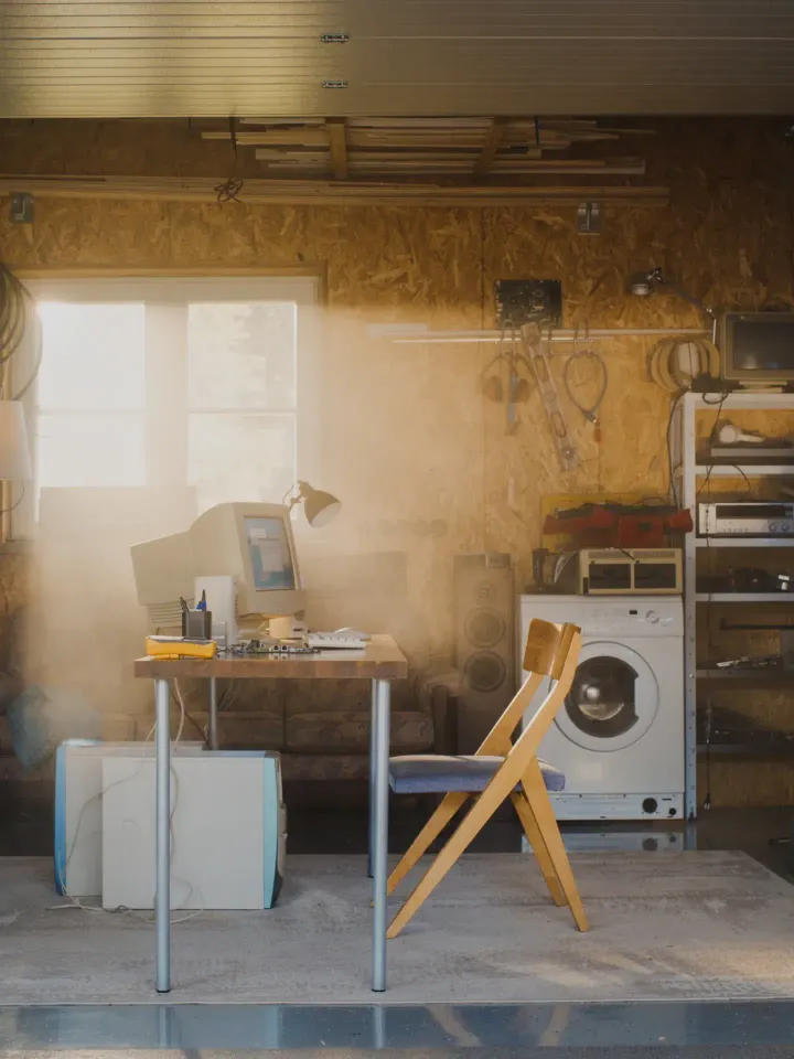 Bureau avec un vieil ordinateur dans un garage avec une fenêtre avec une machine à laver et des étagères avec des bibelots en fond